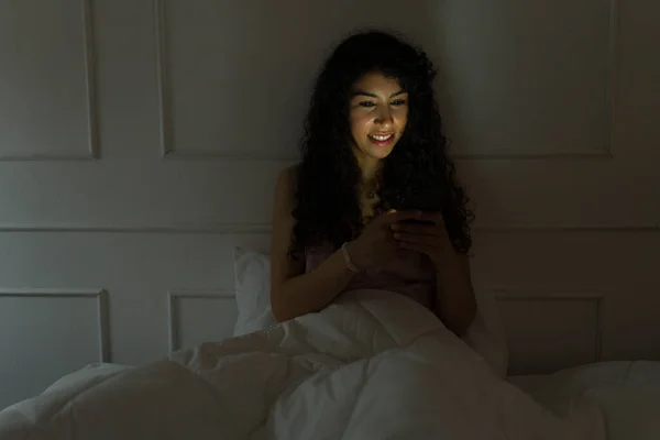 快乐的拉丁裔年轻女性在睡前在智能手机或社交媒体上笑着发短信 — 图库照片