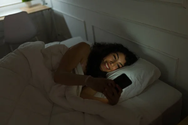 兴奋的年轻女人在睡前躺在被窝里笑着 一边用智能手机发短信 — 图库照片