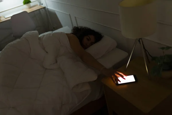 疲惫的年轻女人在床上早上醒来 关掉智能手机上的闹钟 — 图库照片