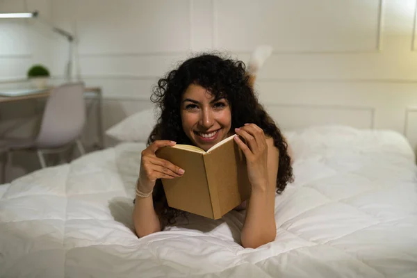 一个美丽的拉丁女人的画像 她一边微笑着 一边眼神交流 一边在床上放松和享受看书的乐趣 — 图库照片