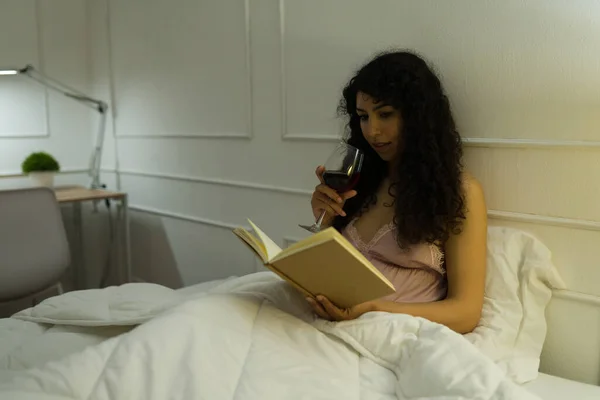 Beautiful latin woman with curly hair drinking wine and reading a book in bed to relax during the night