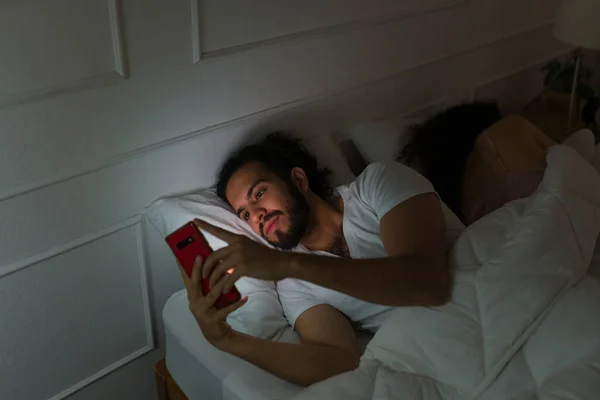 Jovem Bonito Deitado Acordado Noite Sorrindo Enquanto Mensagens Texto Smartphone — Fotografia de Stock