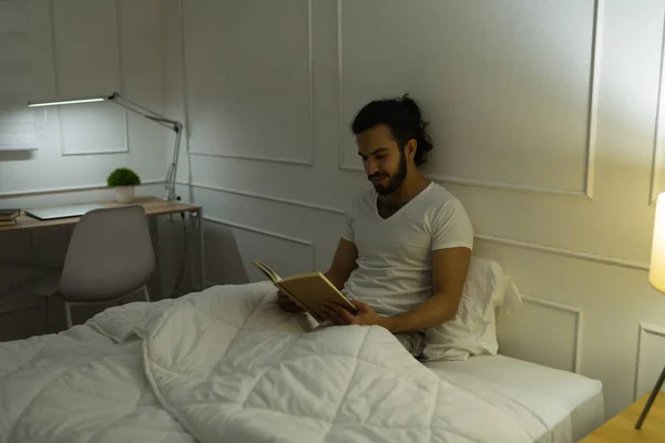 魅力的なリラックスした若いです男に横たわっていますベッドでパジャマで夜に寝る前に面白い本を読んで — ストック写真