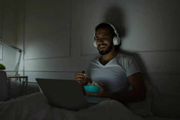 幸せな男笑いながら映画を見ながら食べるポップコーンとともにノートパソコン上でベッドの中で — ストック写真