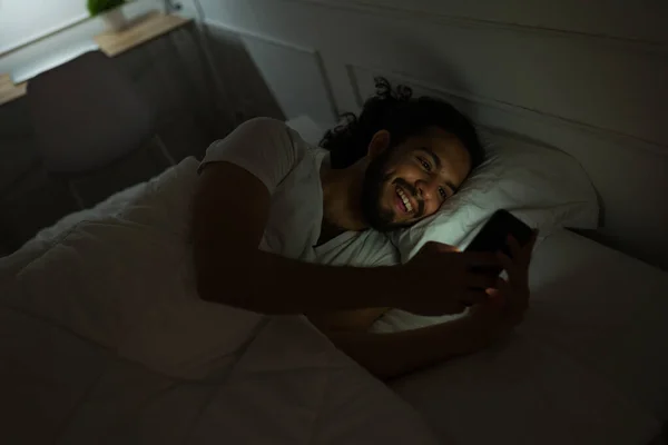 Relaxed happy man smiling texting on the smartphone in the middle of the night before going to sleep
