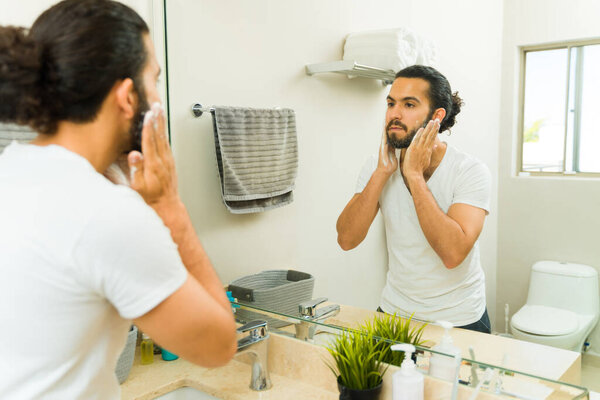 Young man washing his face and beard while using hygiene products looking in the bathroom mirror