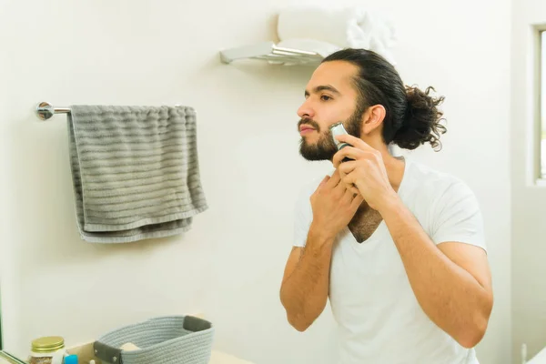 迷人的年轻人在20多岁的时候用修整器在浴室的镜子里修整胡子 — 图库照片