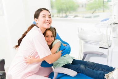 Güzel bayan pediatrik dişçi gülümsüyor. Sevimli bir çocuğa sarılıyor. Diş tedavisinden sonra mutlu görünüyor.