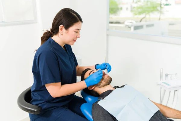 身穿蓝色牙刷的女牙医一边微笑 一边为一名年轻男子做牙科检查 并检查蛀牙情况 — 图库照片