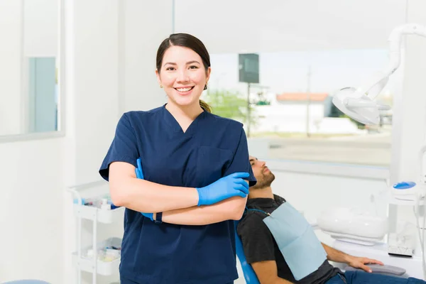 青い布と手袋を身に着けている魅力的な白人女性歯科医患者を持ちながらカメラを見て笑顔 — ストック写真
