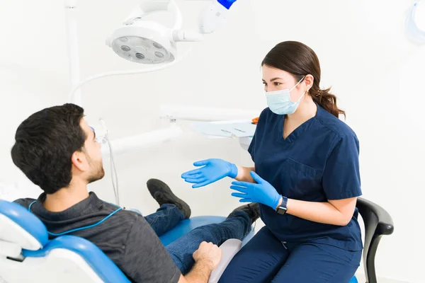 有魅力的女牙医 带着口罩和刷子 向男性病人解释蛀牙的治疗方法 — 图库照片