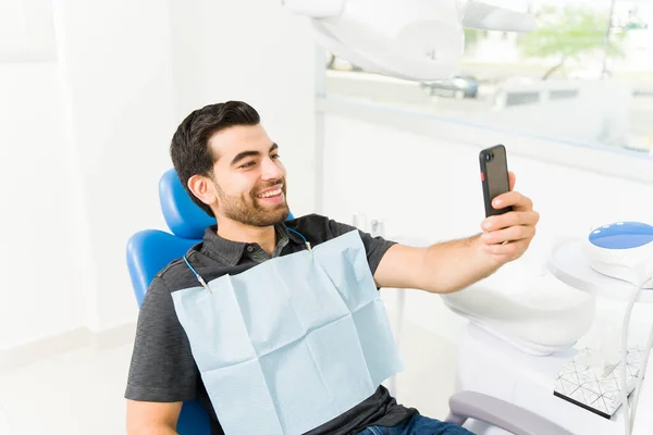 拉丁男人笑得很开心 一边看牙医一边拿着智能手机在社交媒体上发表自己的自拍照 — 图库照片