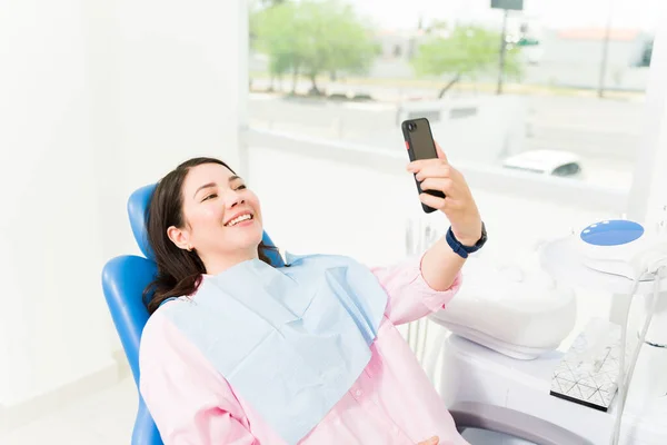白种人女人笑得很开心 一边看牙医一边拿着智能手机 一边在社交媒体上发表自己的照片 — 图库照片