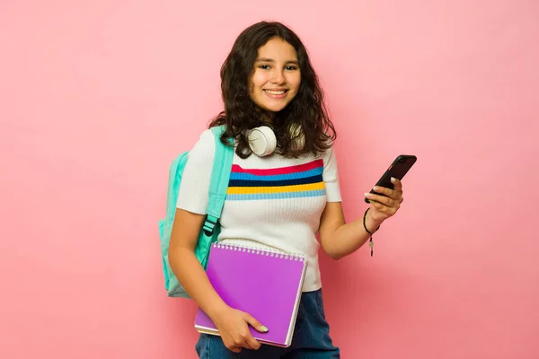 学校でスマートフォンを使いながら ピンクの背景の前でリュックや本を持ちながら笑顔で笑顔を見せる10代の女の子 — ストック写真