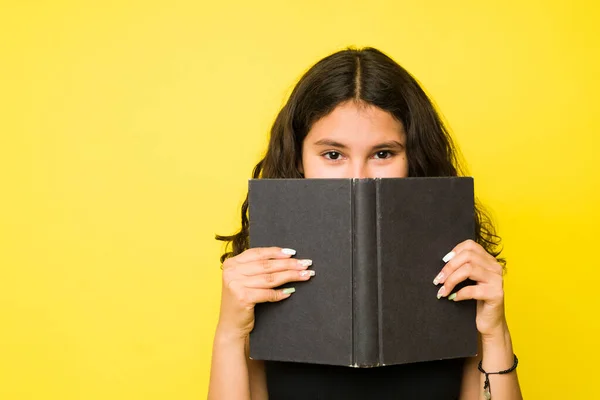 一个可敬的害羞少女躲在一本书后面 在黄色的工作室背景下阅读和欣赏着一本小说 — 图库照片