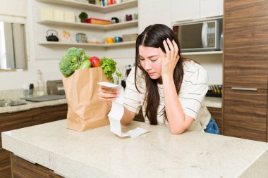 Mutfaktaki endişeli kadın, kötü ekonomide süpermarketten yiyecek aldıktan sonra pahalı fişe bakarken strese giriyor.