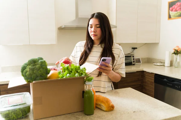 スマートフォンアプリを使ってスーパーマーケットからヘルシーな食べ物をオンラインで注文した後 ベジタリアン女性がキッチンで宅配パッケージを開く — ストック写真