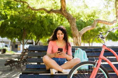 Rahatlamış genç bir kadın parkta bankta oturuyor ve dışarıda bisiklete bindikten sonra akıllı telefonuyla mesajlaşıyor.