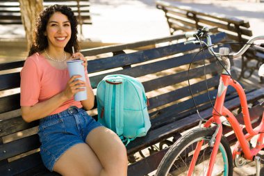 Gülümseyen genç bir kadın mutlu bir şekilde göz teması kuruyor yazın tadını çıkarıyor bisikletinin üstünde oturmuş kahve içiyor.