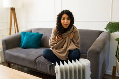 Soğuk genç bir kadın, elektrikli ısıtıcının önünde battaniyeye sarılı otururken kameraya bakıp titriyor ve kötü hissediyor.