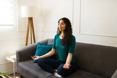 Rahat genç bir kadın, meditasyon müziği dinliyor, yoga yapıyor ya da evde nefes egzersizleri yapıyor. Kanepede oturuyor.