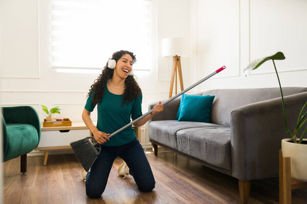 Excited young woman doing housework and cleaning chores while having fun and sweeping the living room listening to music