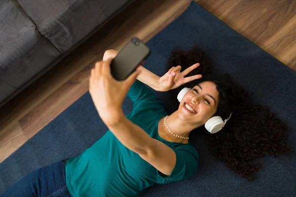 床の上でリラックスしながら ヘッドフォンでスマートフォン上でオンラインビデオ通話を持つ魅力的なヒスパニック系女性の高角度 — ストック写真