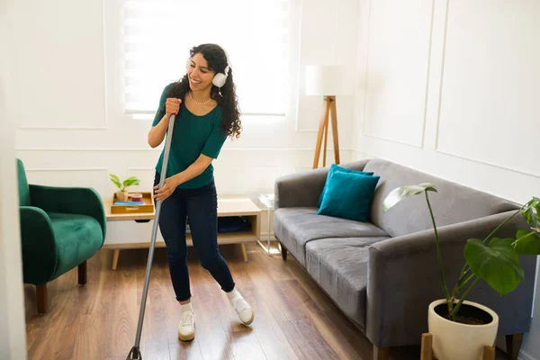 ヘッドフォンで音楽を聴きながら 掃除の雑用をしながら 床を掃除している陽気な若い女性 — ストック写真