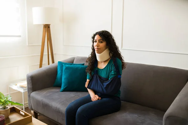 伤心受伤的年轻女子一个人坐在沙发上 胳膊断了 脖子上的支撑架在家里感觉很糟 — 图库照片