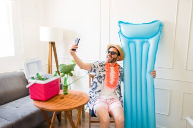 30 'lu yaşlarındaki mutlu Latin adam şişirilebilir bir fotoğraf çekerken ve sahildeymiş gibi davranırken ve evde kalıyormuş gibi yaparken.