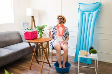 Relaxed happy man smiling while having a pedicure at home while using a fan during the summer vacations wearing shorts and sunglasses clipart