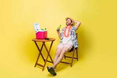 40 'lı yaşlarda, plajdaki yaz tatili boyunca oturup bira içen ve dinlenen rahat beyaz bir adam sarı bir arka plana yaslanmış.