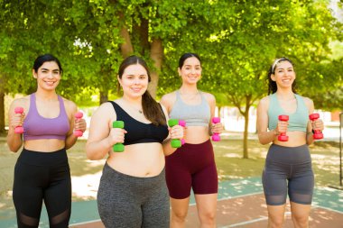 Gülümseyen, göz teması kuran ve vücut çeşitliliğini kutlarken halter ağırlıklarıyla egzersiz yapan neşeli bir grup kadın arkadaş.