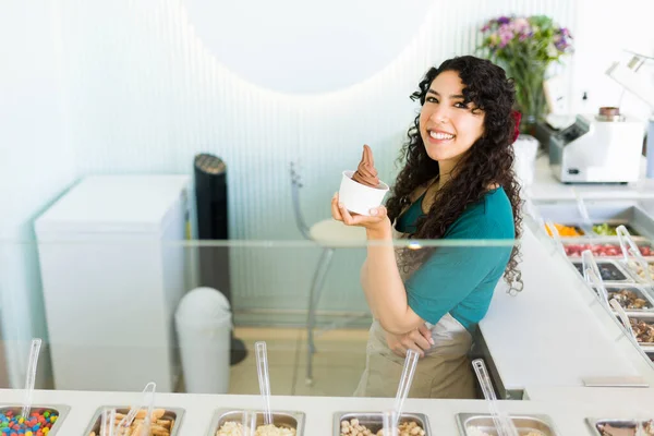 在冰淇淋店工作的漂亮而快乐的女人 吃着美味的巧克力冰淇淋 面带微笑地进行眼神交流 — 图库照片