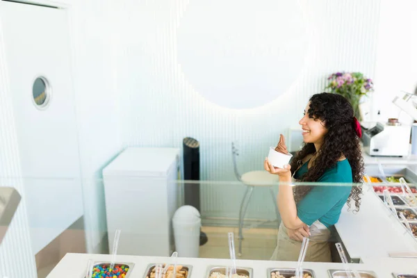 在冰淇淋店的顶级吧台后面 一位快乐的年轻女士正坐在餐厅的柜台后面 一边吃着上面涂满糖霜的酸奶 一边享受着美食 — 图库照片