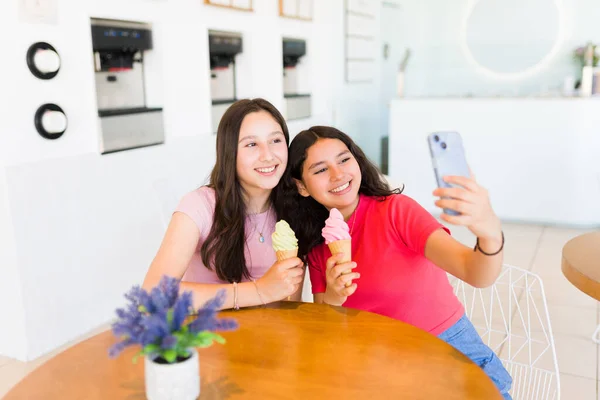 可爱的少女们一边在冰淇淋店玩乐 一边在智能手机上自私自利 吃着冰冻的酸奶蛋筒 — 图库照片
