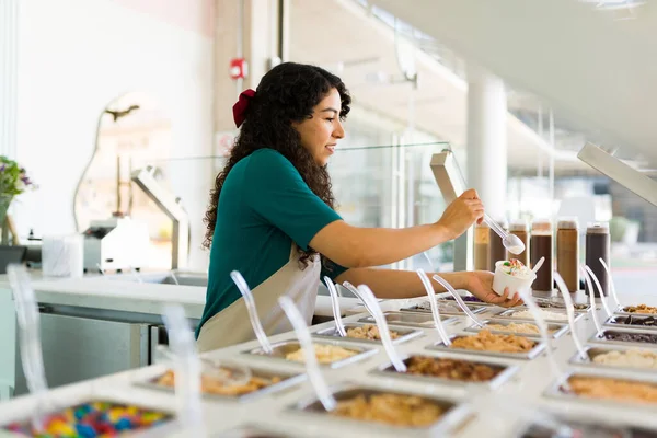 年轻女工一边在商店柜台后面工作 一边笑着准备美味的冷冻酸奶和配料 — 图库照片