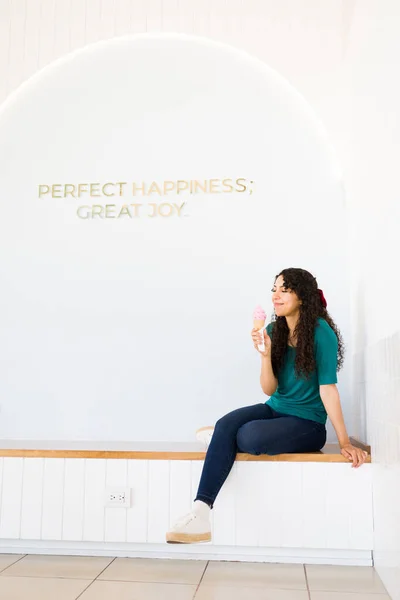Relaxed happy woman enjoying eating an ice cream cone at the frozen yogurt shop and sitting in front of a white wall with copy space
