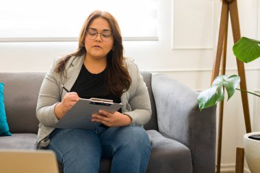 Gözlüklü profesyonel kadın psikolog. Pano kullanıyor ve terapi sırasında hasta değerlendirmesi yaparken not alıyor.