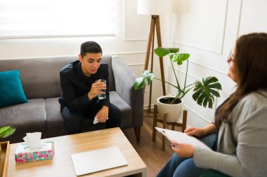 Üzgün bir adamın kederli bir şekilde bir bardak su içmesi ve bir psikologla yas tutarken ağlaması.