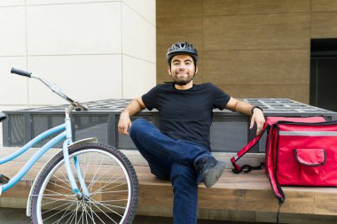 Rahatlamış teslimat işçisi şehirde oturmuş sırt çantası ve çevre dostu bisikletiyle teslimat yapmayı bekliyor.