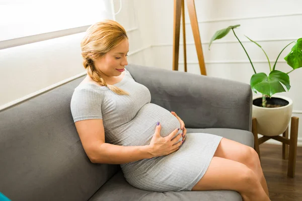 可愛いです妊娠中の女性の肖像画上のソファに家に座ってと彼女の腹に触れる — ストック写真