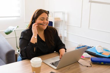 Güzel şişman satış temsilcisi müşteriyle konuşuyor kulaklık takıyor ve ofisteki laptopta gülümsüyor.