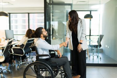 Engelli iş adamı tekerlekli sandalyede oturmuş modern ofiste Asyalı iş kadınıyla el sıkışıyor.
