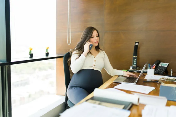 Embarazada Empleada Sentada Escritorio Hablando Teléfono Inteligente Mientras Usa Computadora — Foto de Stock