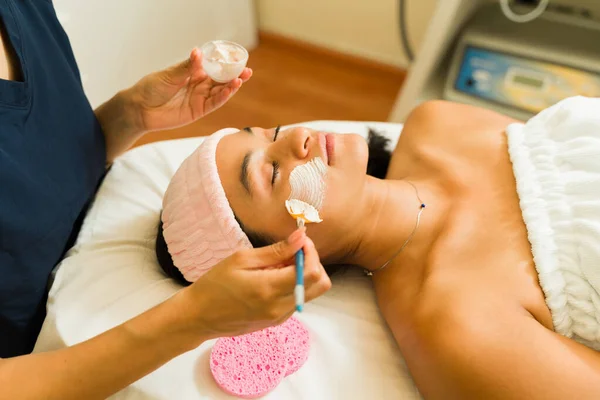 Relaxed hispanic woman getting a facial mask and exfoliation treatment at the beauty spa while relaxing and resting
