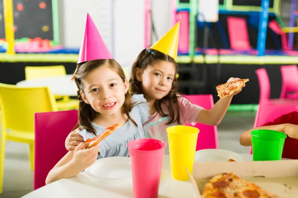 Adorable little girls and friends hugging while wearing party hats and eating pizza with cake celebrating a friends birthday