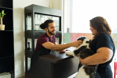 Güzel bir köpeği okşayan ve kuaförde saç tıraşı için shih tzu getiren bir kadınla konuşan mutlu bir kuaför.