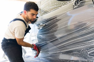 Araba yıkama eldiveniyle araba yıkama servisinde köpük uygulayan profesyonel genç bir çalışan.