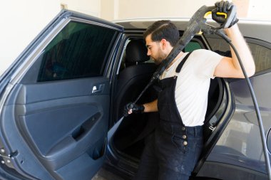 Genç Latin erkek işçi araba yıkamada basınçlı su hortumuyla bir aracı yıkıyor.